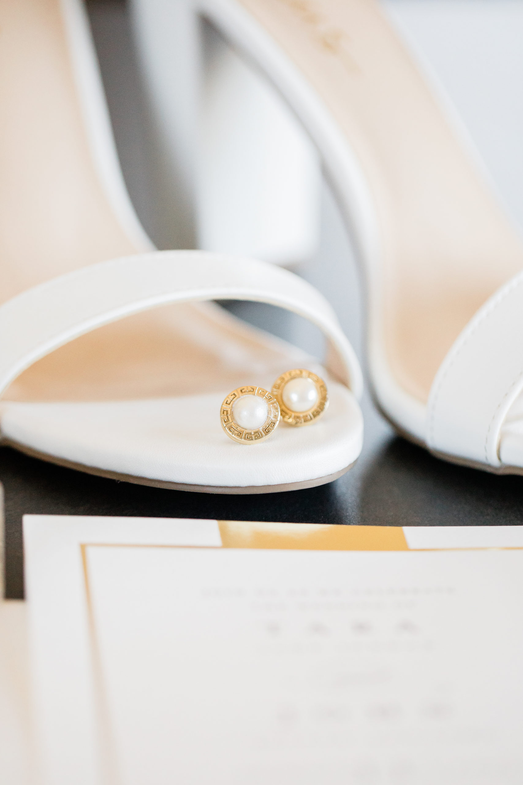 A pair of gold and pearl earrings placed on the toe of a white high heeled shoe.