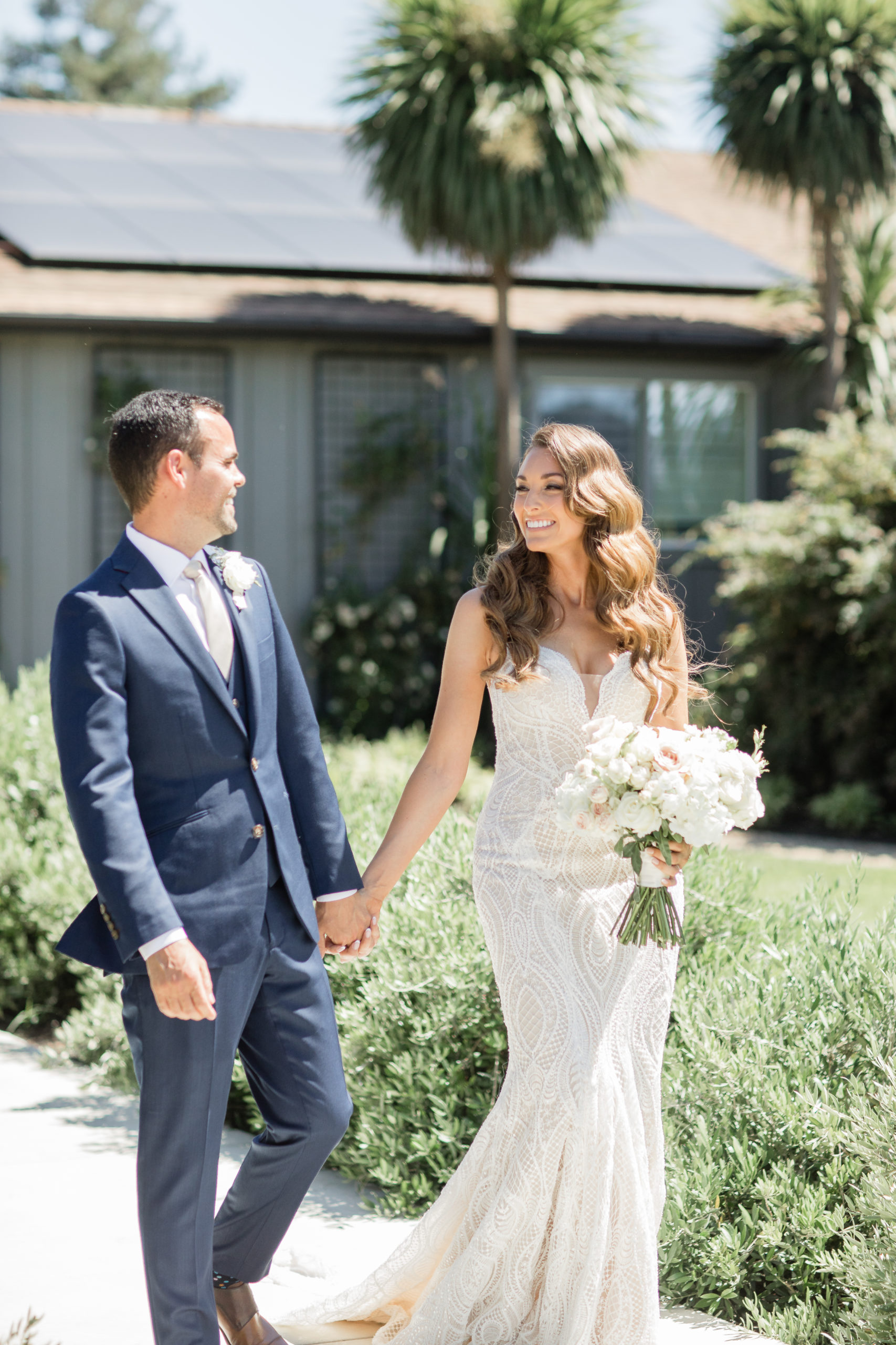 Tara and John in wedding attire, looking at each other and smiling as they walk down a concrete path with a house and two palm trees in the background.