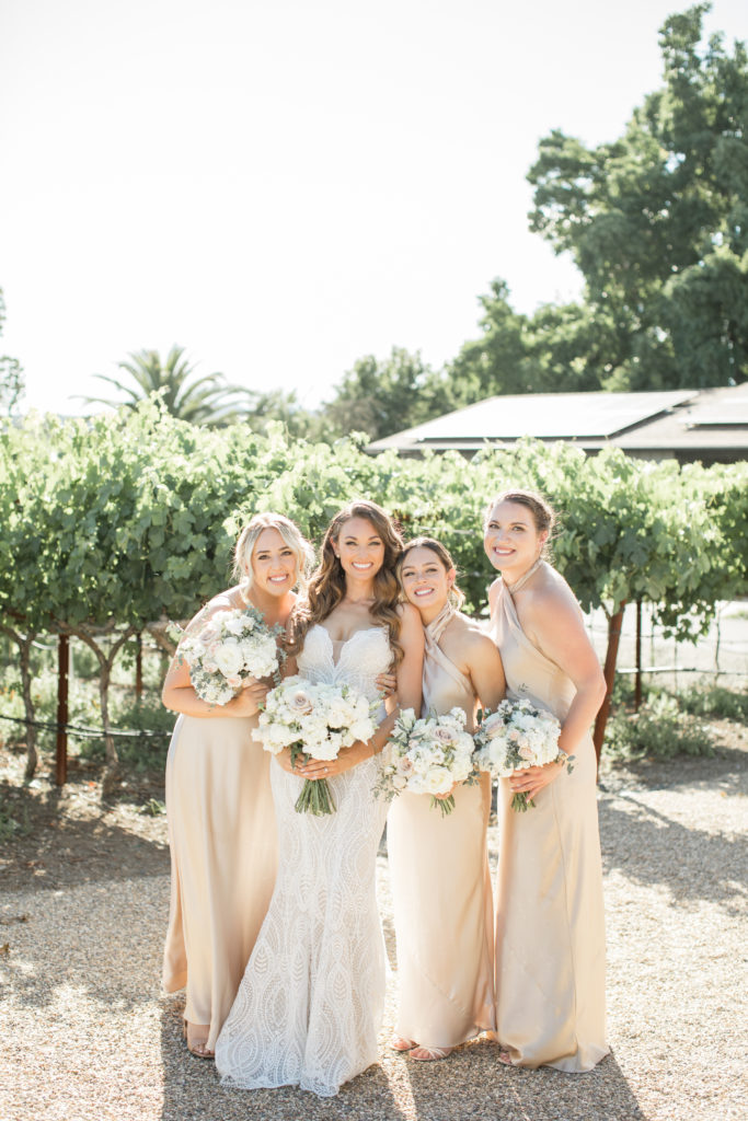 Tara in her wedding dress with three bridesmaids in champagne colored dresses with vineyards in the background, smiling looking at the camera.