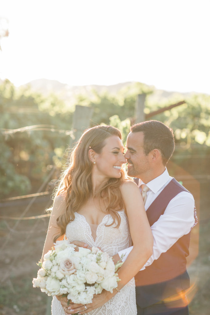 A newlywed couple looking into each others' eyes, smiling with vineyards in the background at golden hour.
