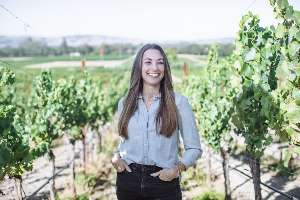 Fun fact 5 is a girl with brown hair in blue button up shirt smiling at the camera with vineyards in the background.