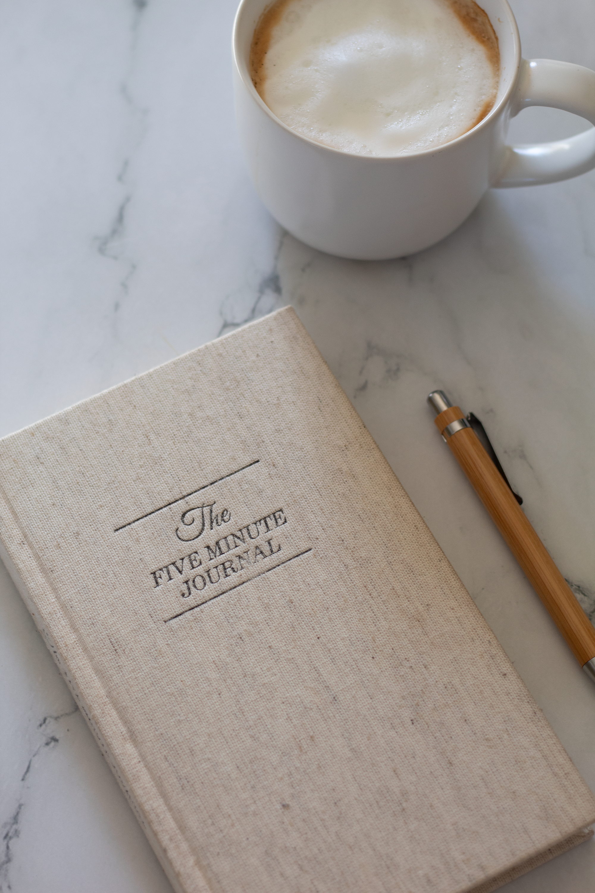 The linen colored cover of The 5 Minute Journal by Intelligent change sitting on a marble counter with a wood pen on the right and a coffee in a white cup above it.