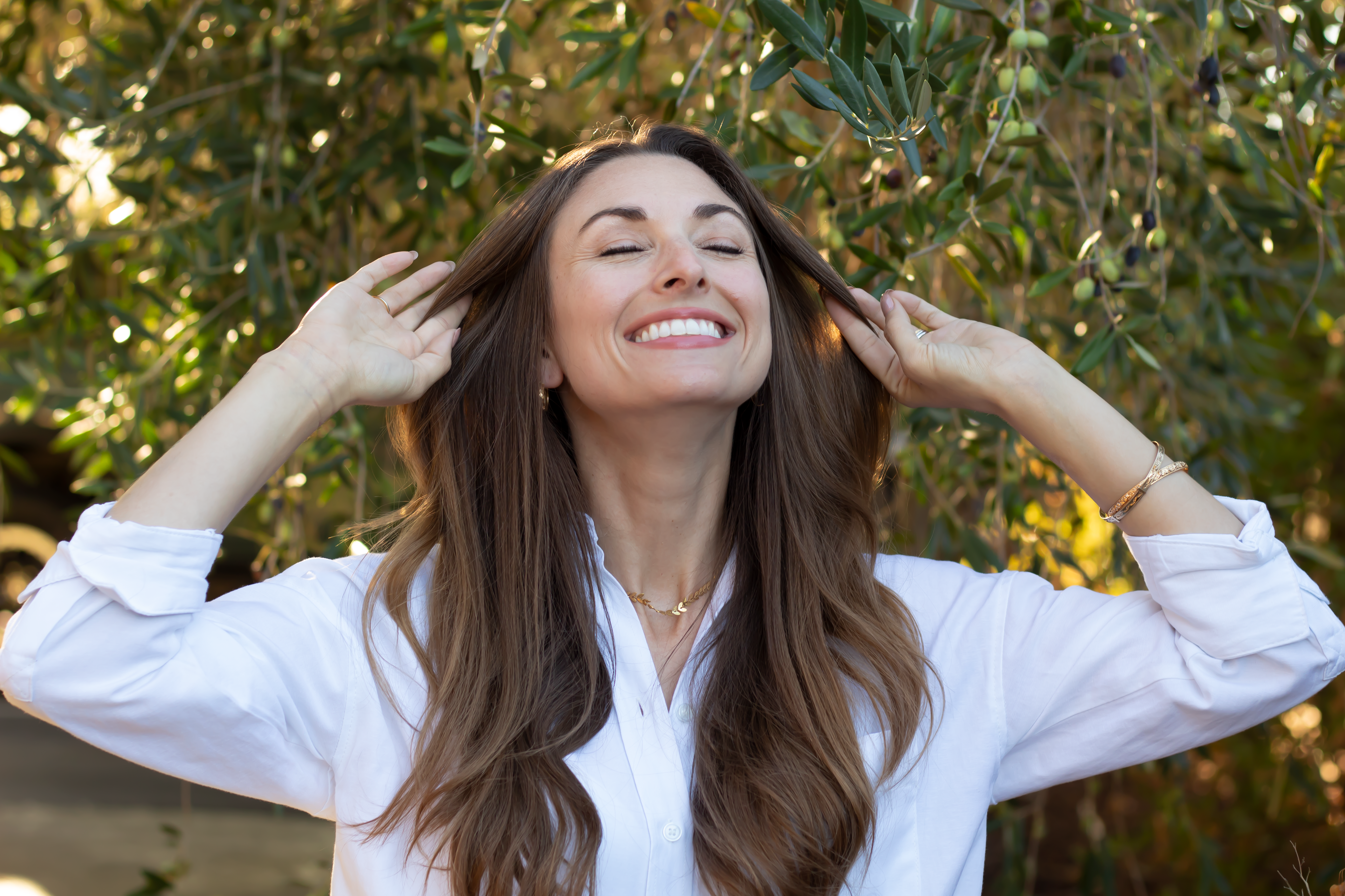 Woman with long brown hair in a white button up shirt smiling and looking up with her eyes closed