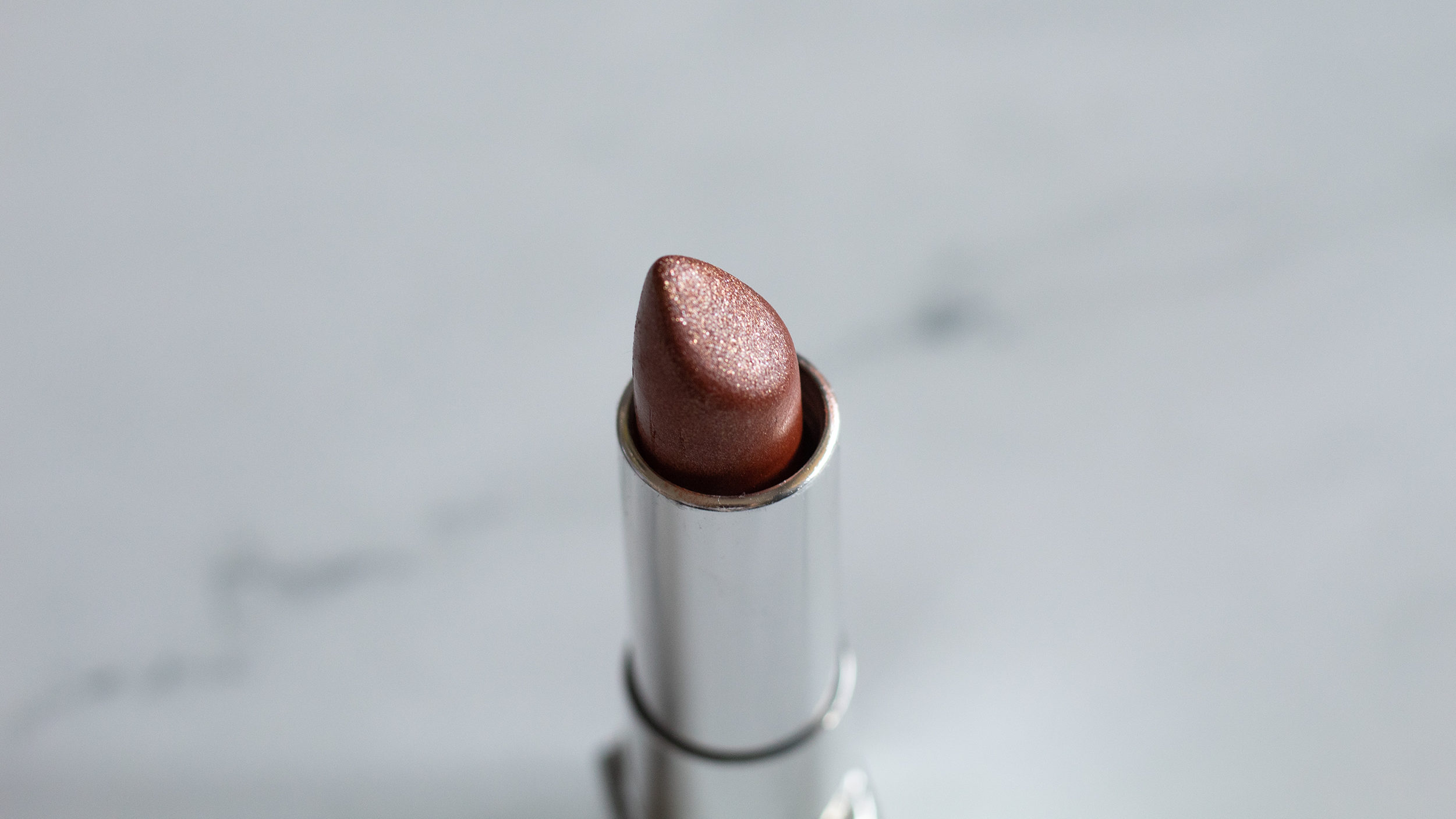 A silver tube of lipstick with a copper/gold lipstick against a white marble background