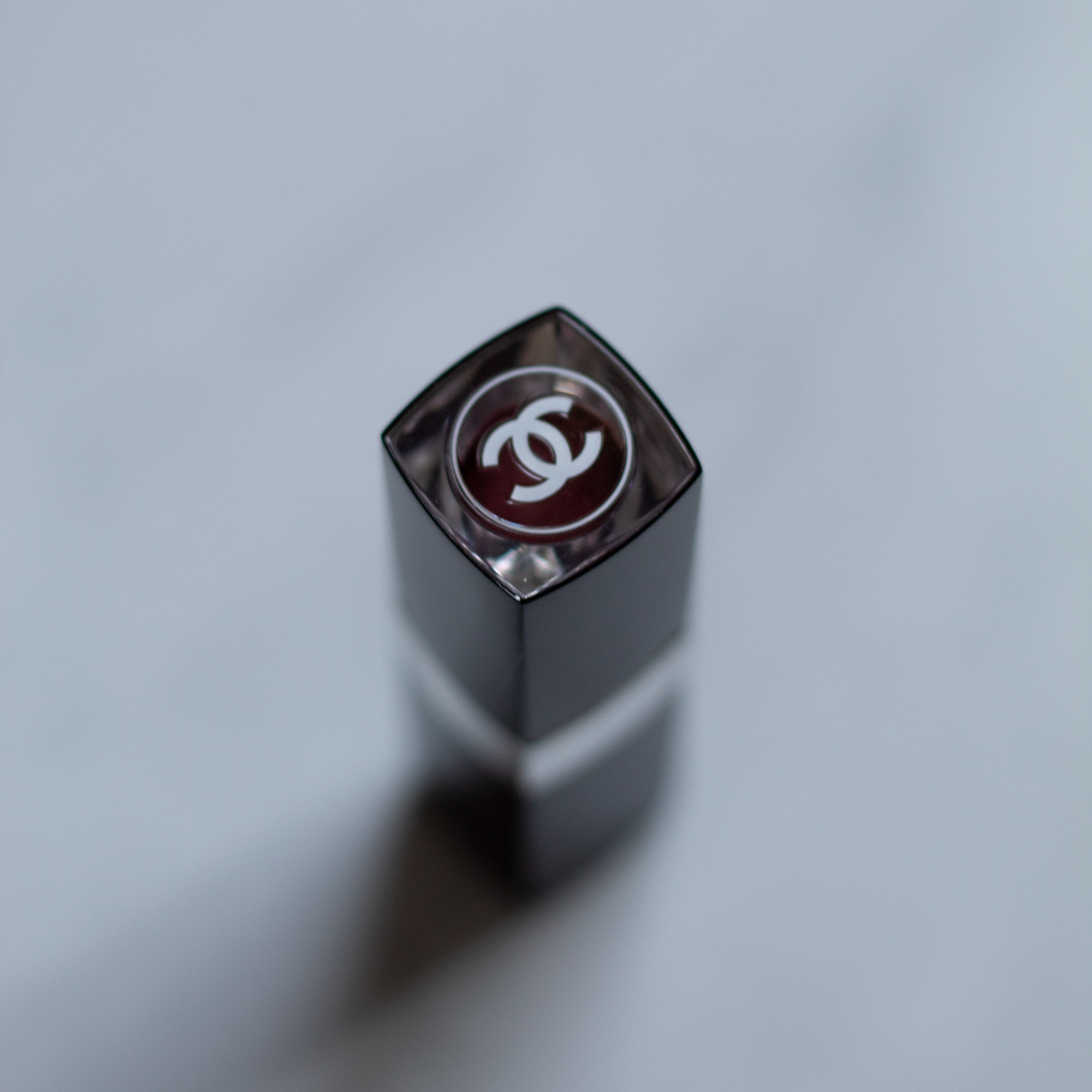 A black and silver lipstick tube standing up against a white marble background and the Chanel double C on top