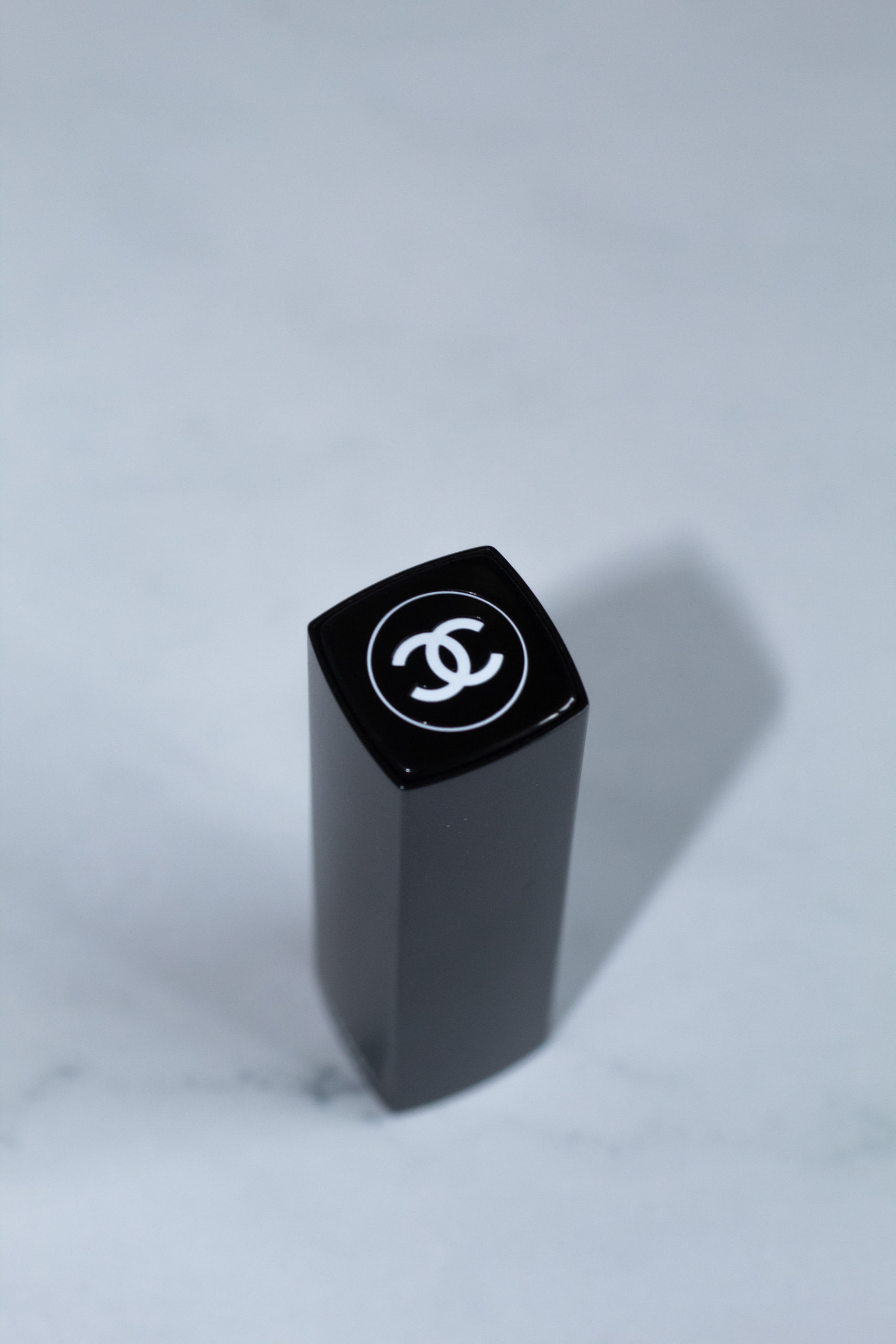A black lipstick tube with the white Chanel double C on top against a white marble background