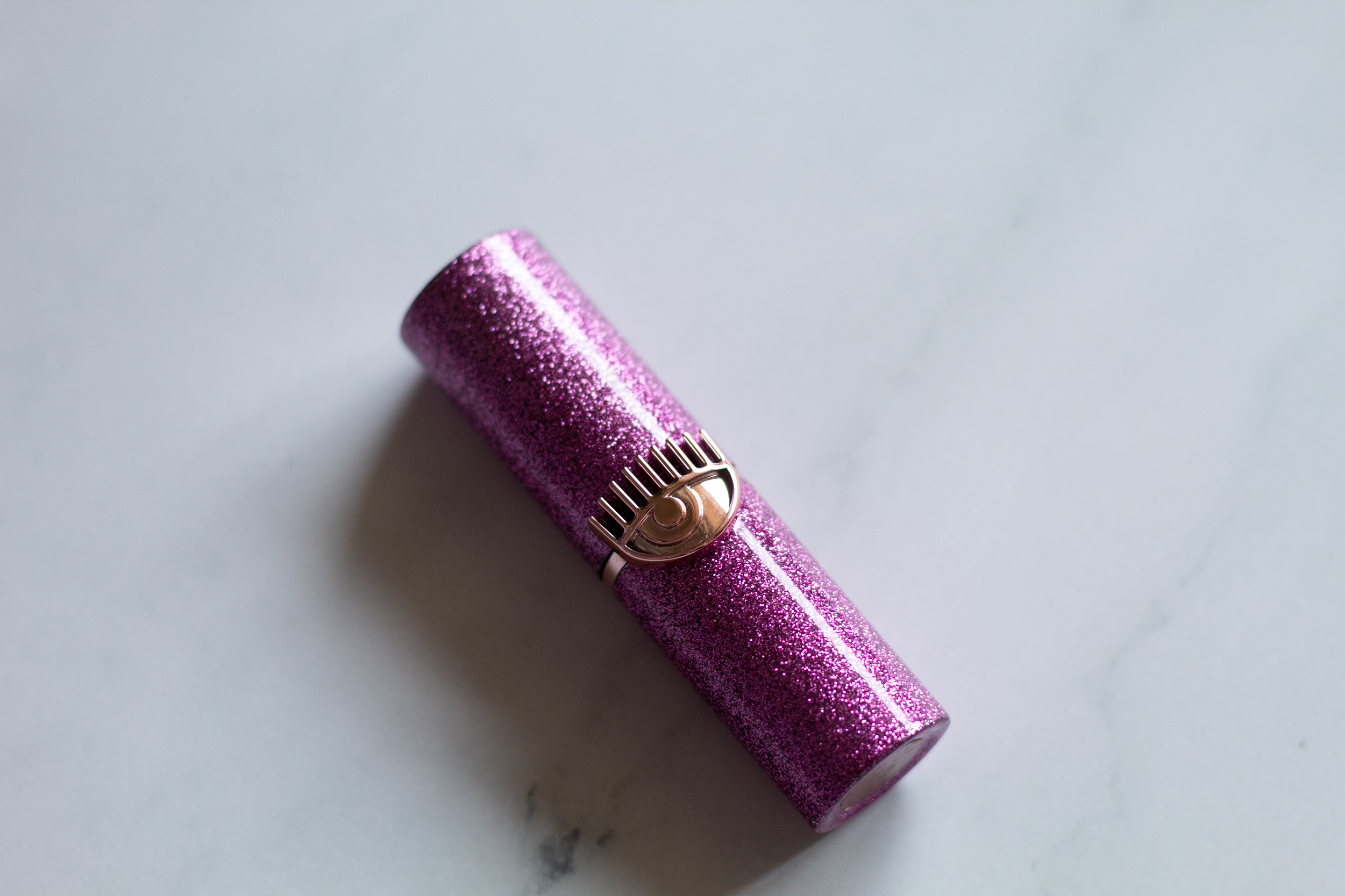 A purple sparkly lipstick rube with a gold eye icon in the middle against a white marble background
