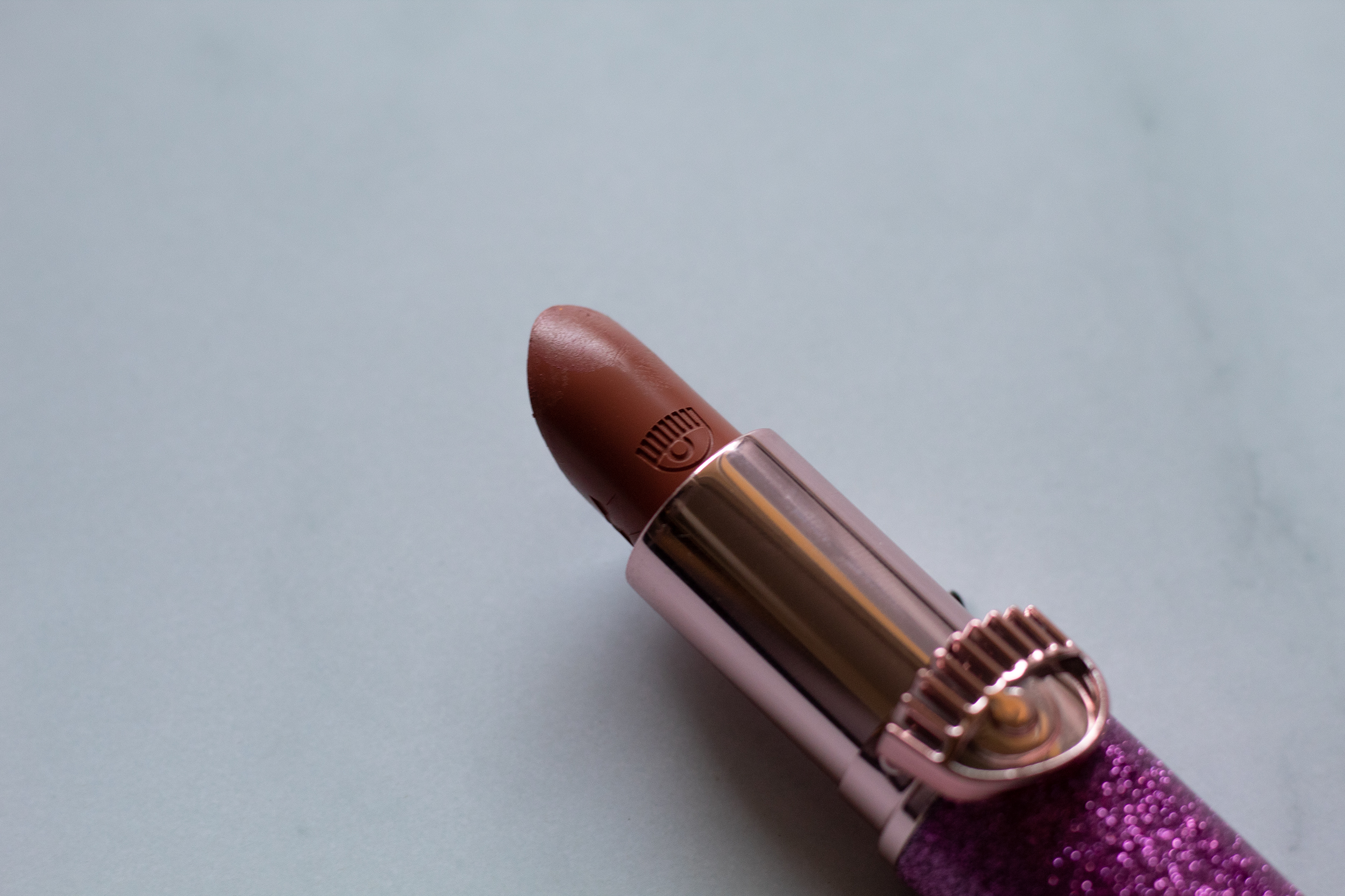 A lipstick in a purple sparkly and gold tube that is open to show a brown rose hue against white marble background