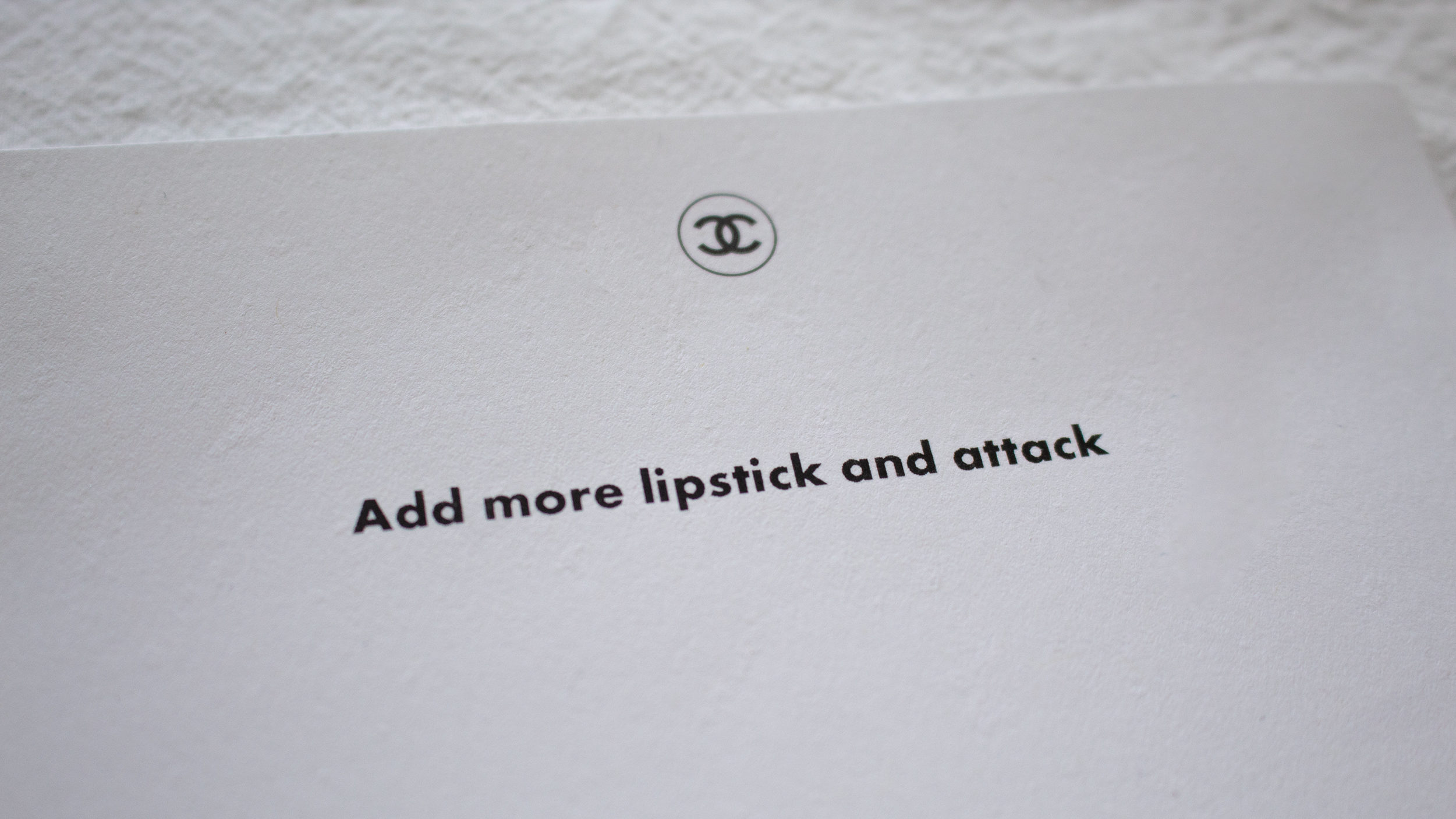 A white card that says Add more lipstick and attack against a white bed comforter