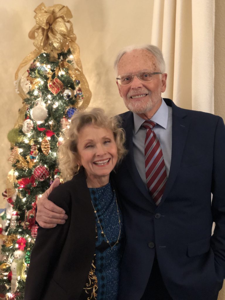 A man and woman couple in their 60's, smiling at the camera with a Christmas tree in the background to honor the holiday