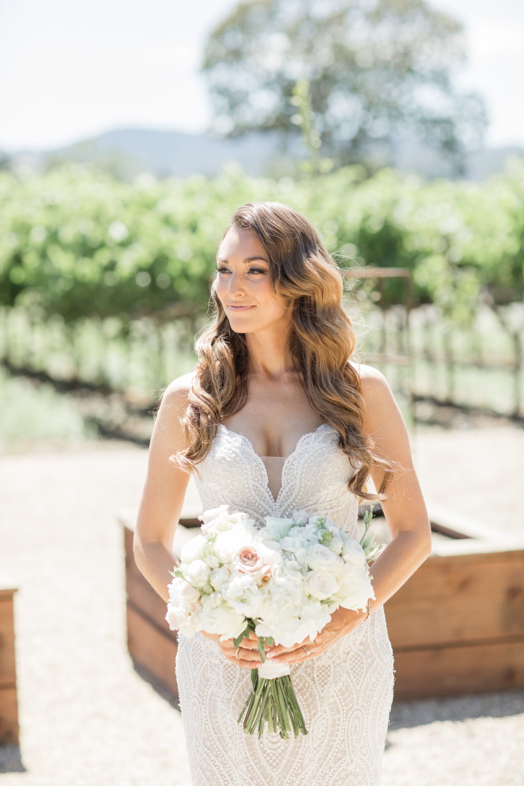 Tara outside in front of a vineyard in her wedding dress and veil, holding her bouquet and looking to the right and smiling.
