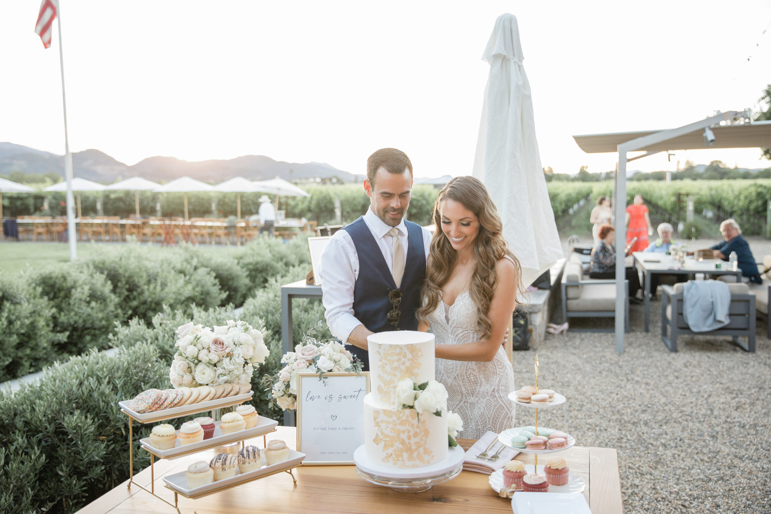 Tara and John cutting their wedding cake at a dessert table with bushes and vineyards in the background.