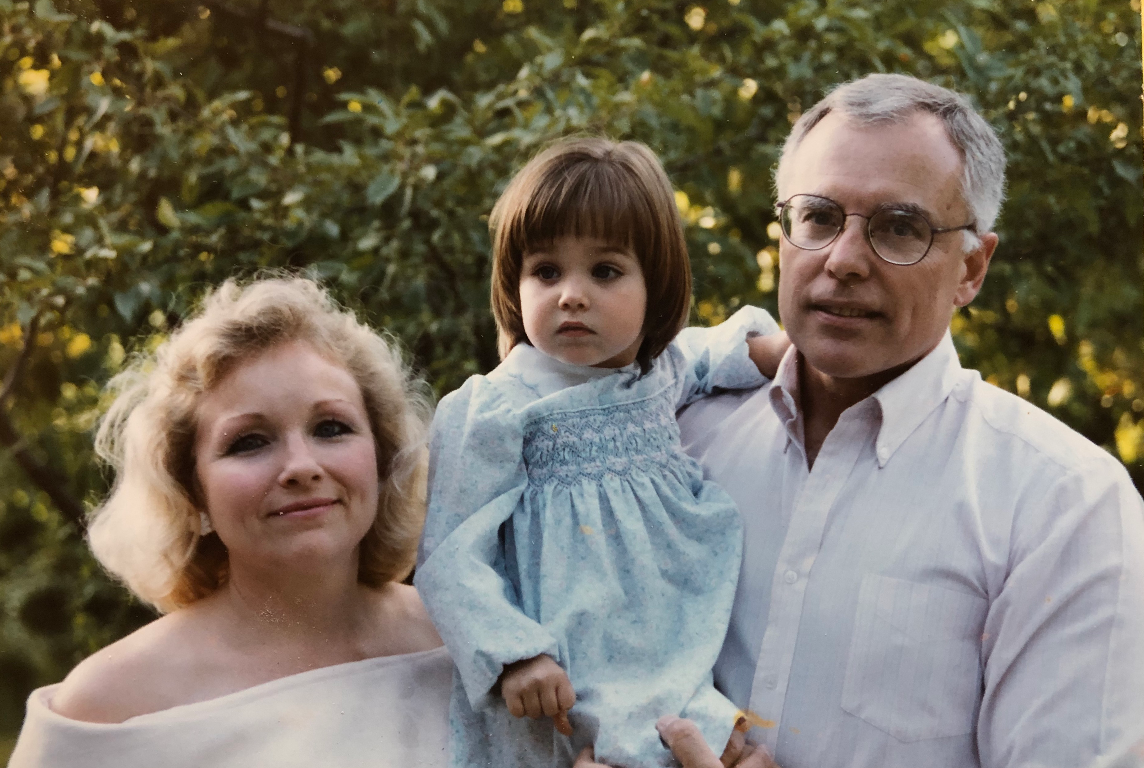 A photo of a young mother, infant girl, and father posing for a photo with greenery in the background