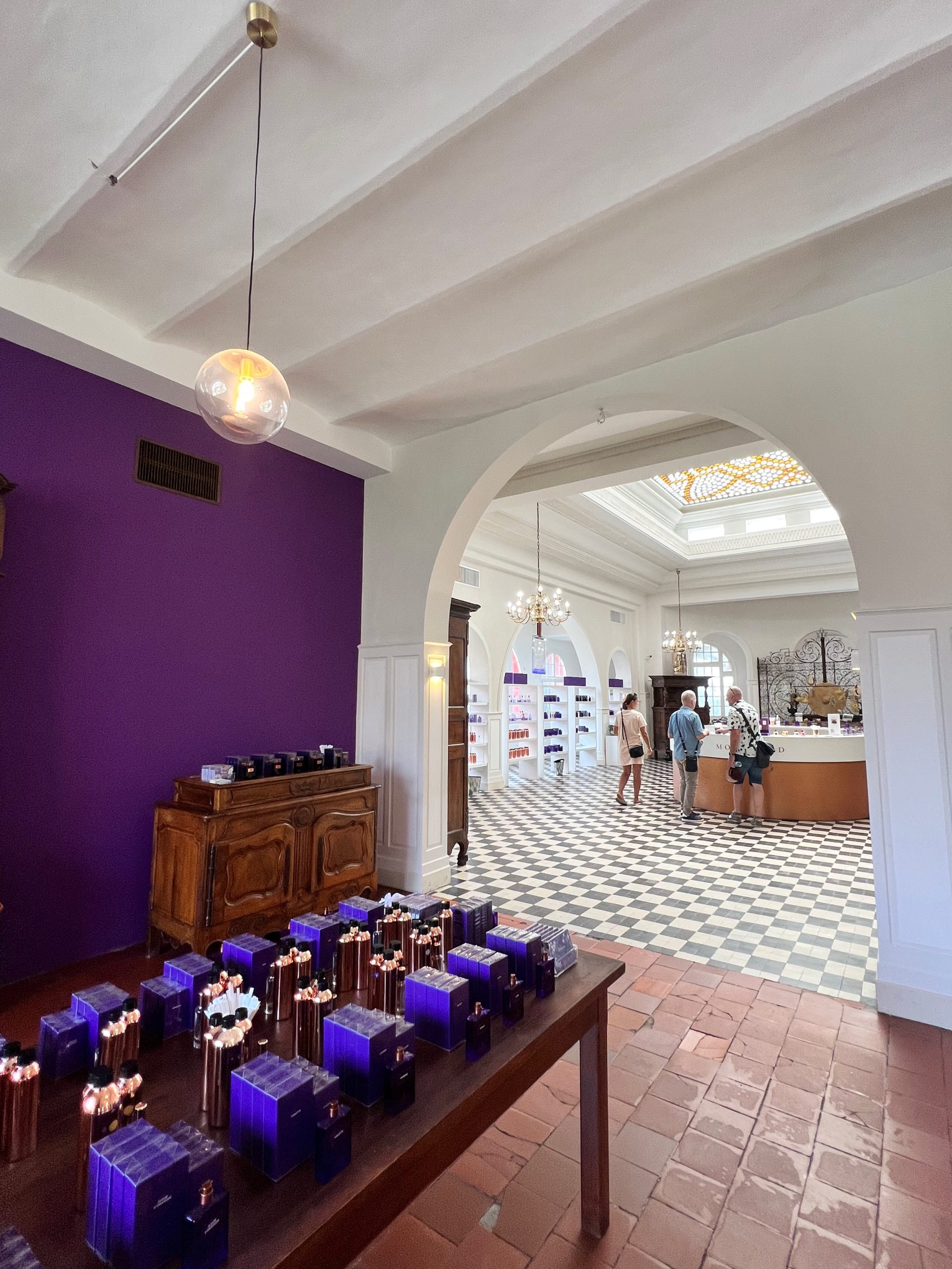 A purple wall on the left with a table with purple perfume boxes in front of it, which opens up to a bright showroom on the right
