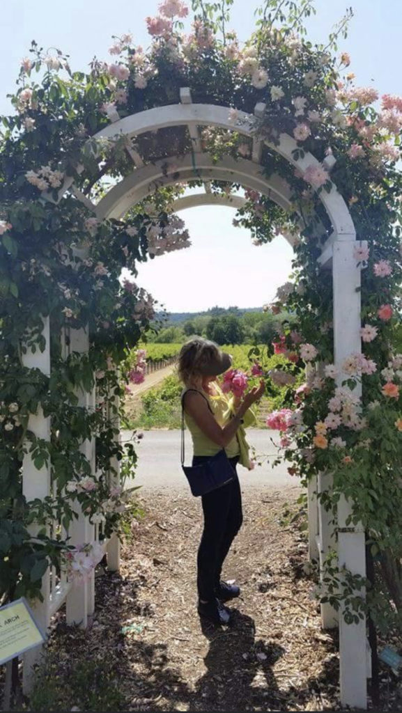 Petite woman with blonde hair under an arch with flowers and greenery, smelling a rose