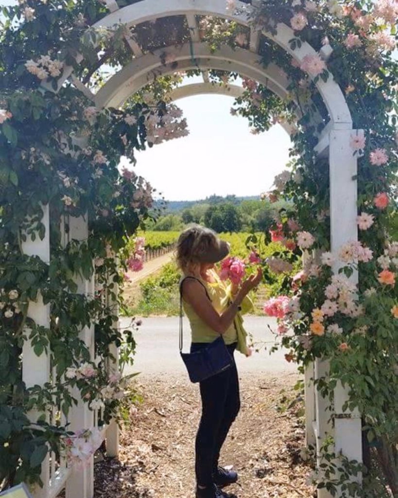 A petite blonde woman underneath an archway of flowers, smelling a rose