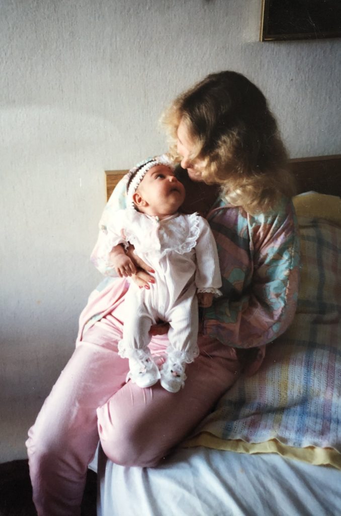 A mother with blonde hair sitting on a bed with a brown-haired baby girl sitting on her lap, looking at each other