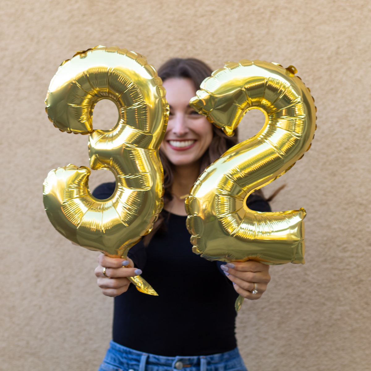 Girl smiling with brown hair and a black shirt holding two gold balloons in the numbers three and two