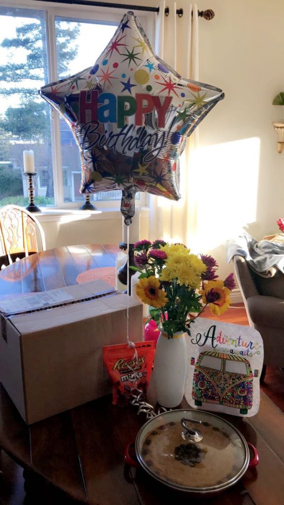 A Happy Birthday balloon with flowers, a brown box, and a chocolate chip cookie cake on a table.