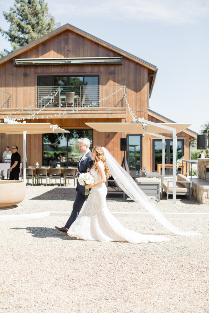 A bride with brown hair walking across light graven with a tall man linking arms in front of a wood building.