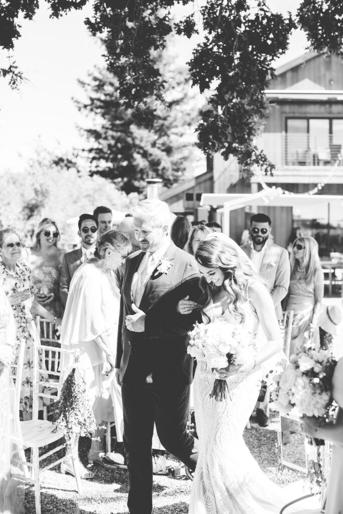 A black and white photo of a bride walking down the aisle of a wedding with a man in a suit.
