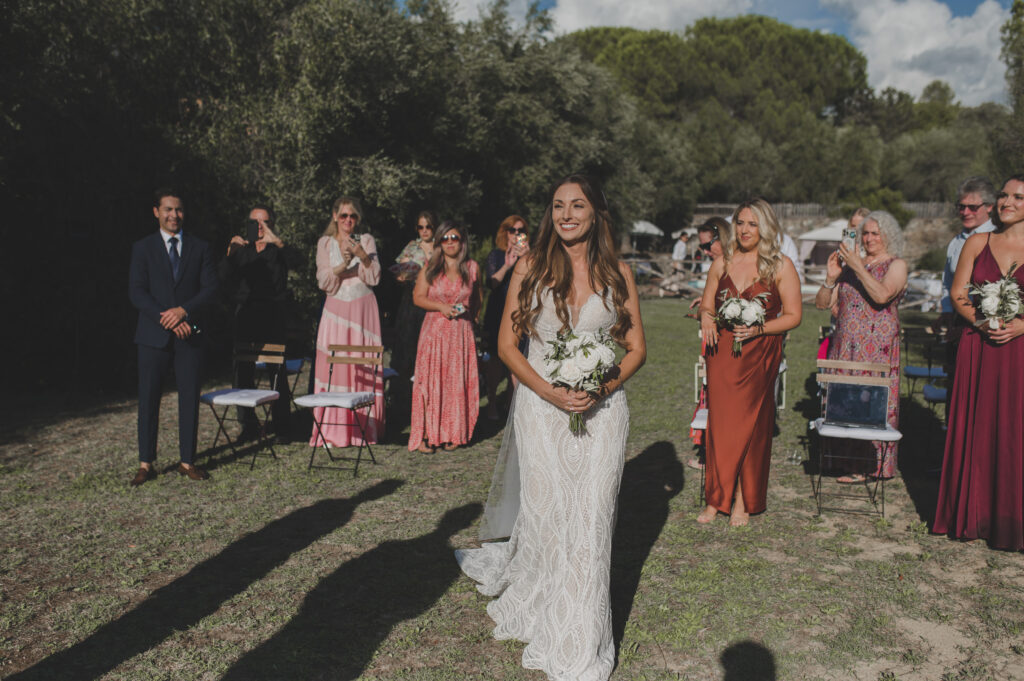 A woman with brown hair in a wedding dress walking down the aisle in an outdoor ceremony.