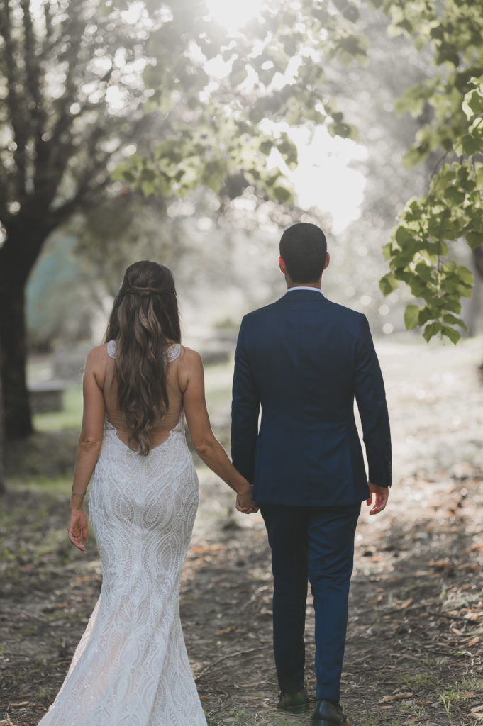 A man with black hair and facial hair in a blue suit walking with a brunette woman away from the camera in a wedding dress.