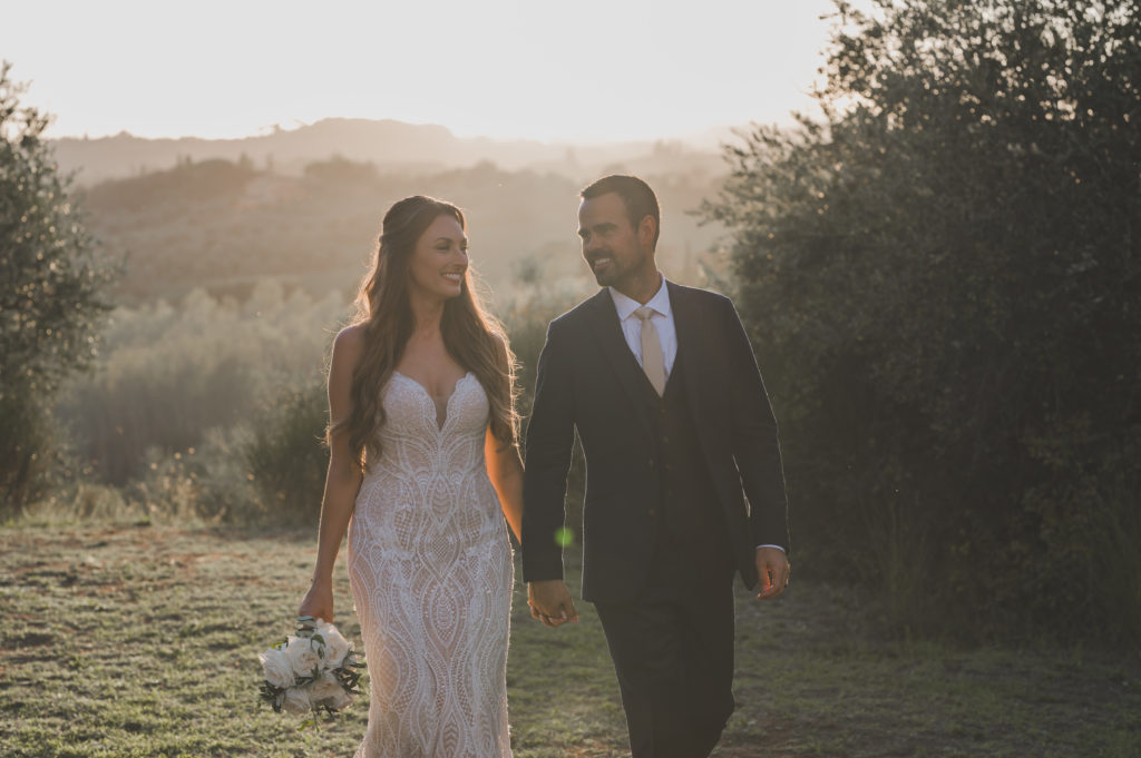 A man with black hair and facial hair in a blue suit walking with a brunette woman in a wedding dress with golden hour hills in the background.