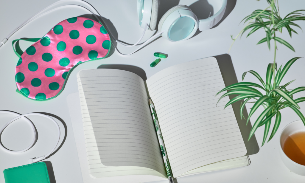 A notebook with white and green headphones, pink and green polka dot eye mask, a green spider plant, and a cup of tea.